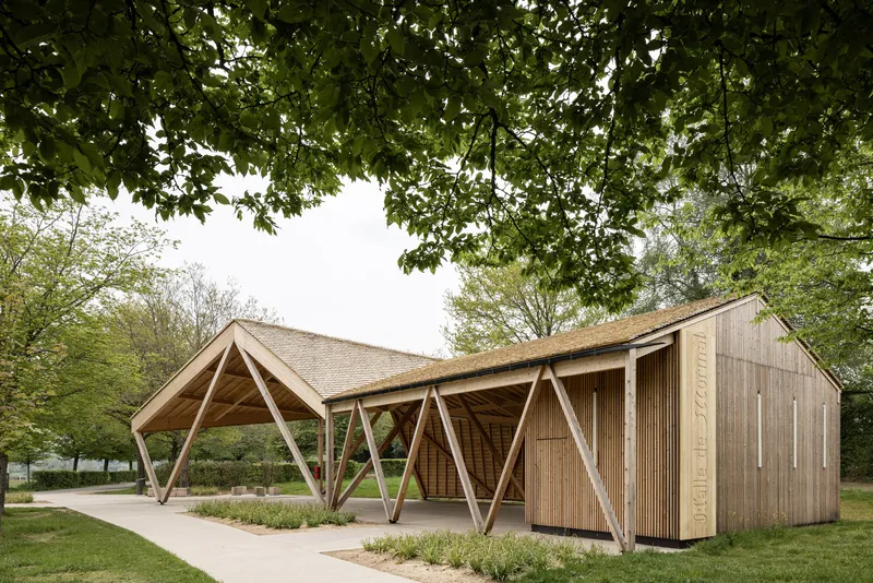 Timber pavilion with diagonal braced columns and vertical wood slat cladding framed by leafy trees overhead