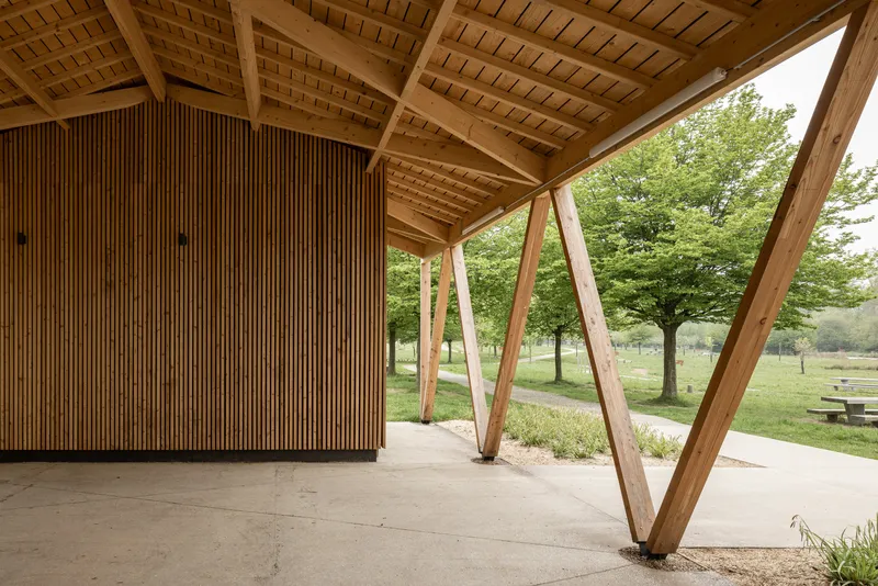 Covered pavilion with angled timber columns and slatted wood wall overlooking a park in summer