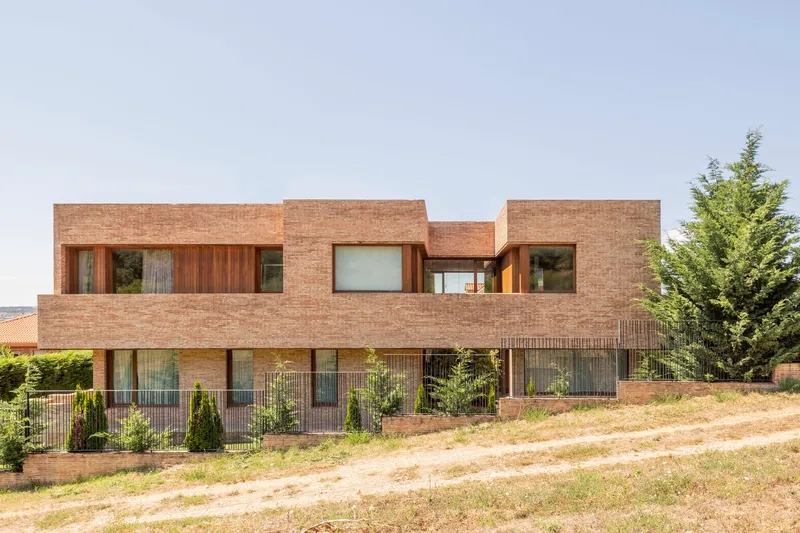 Two-story brick facade with recessed timber-framed windows set above a sloping meadow and wire fence