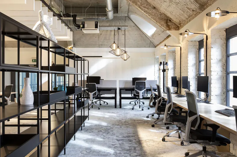 Workspace with desk rows and black shelving lit by pendant fixtures beneath diagonal concrete beams
