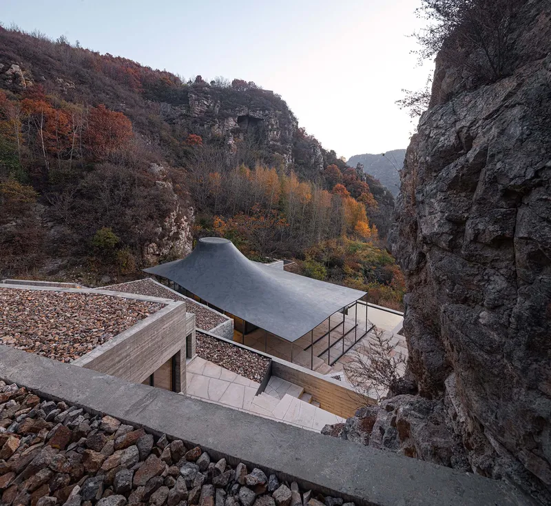 Pavilion with sweeping metal roof set within a narrow gorge framed by autumn foliage at dusk