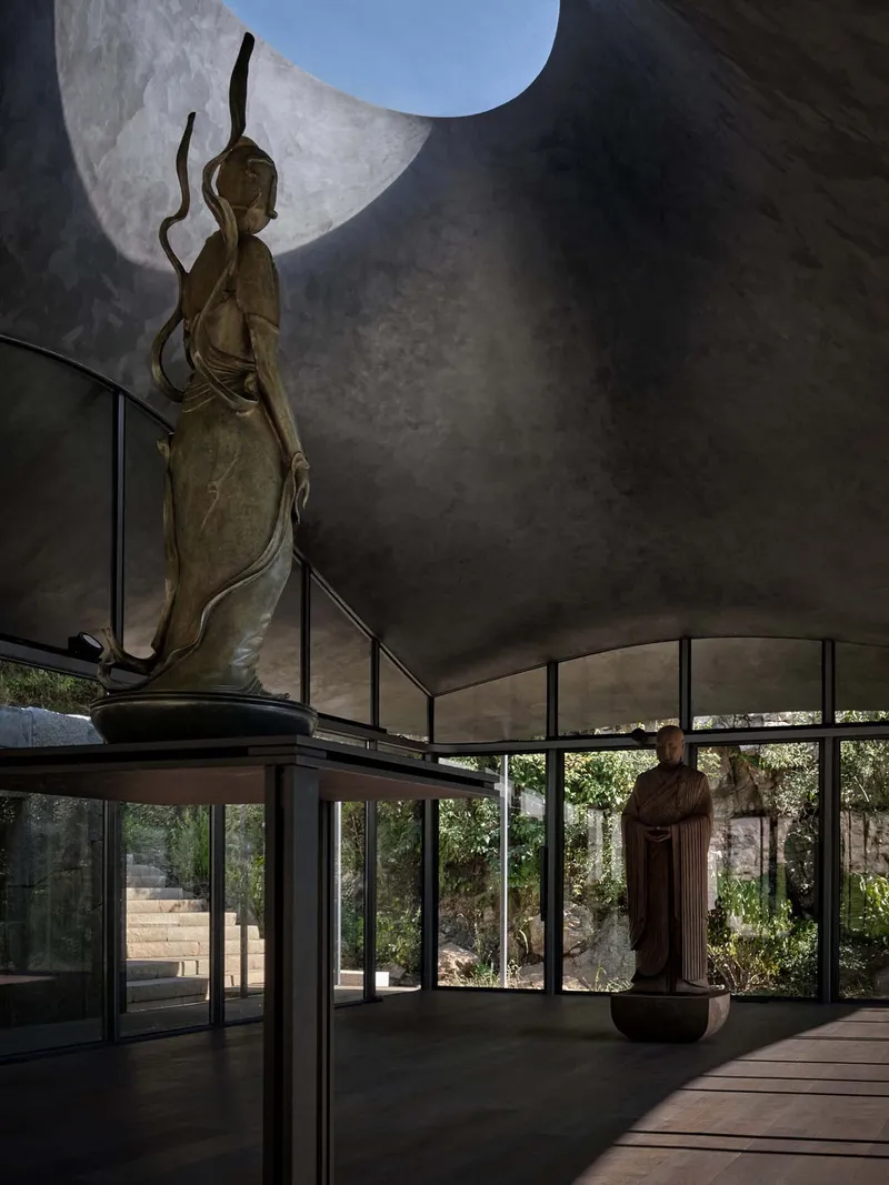 Gallery space with curved ceiling oculus above a bronze statue and glazed walls overlooking the landscape