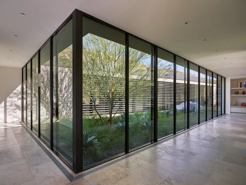 Corner interior courtyard wrapped by full-height black-framed glazing overlooking a planted garden with a young tree