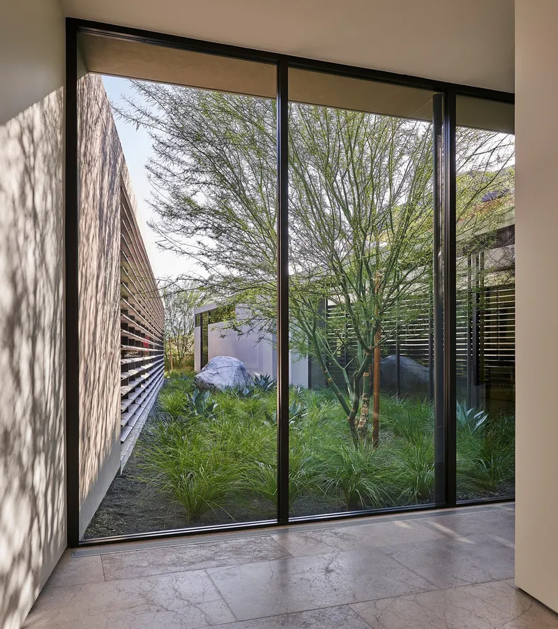 Floor-to-ceiling window framing an enclosed courtyard with native grasses and a sculptural boulder under dappled shadow