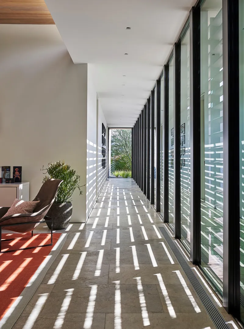 Long hallway with louvered screen wall casting striped sunlight across limestone floor and timber ceiling insert