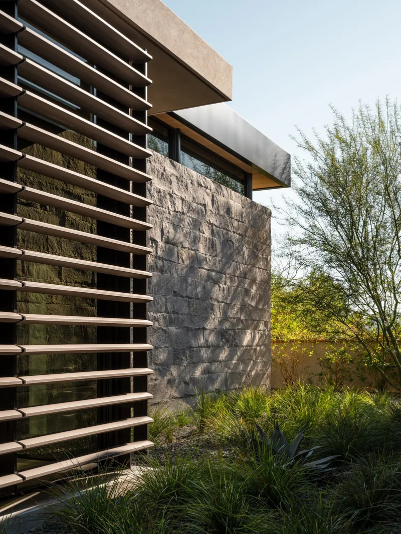 Stone wall beside horizontal timber louver screen and planted bed with ornamental grasses at twilight