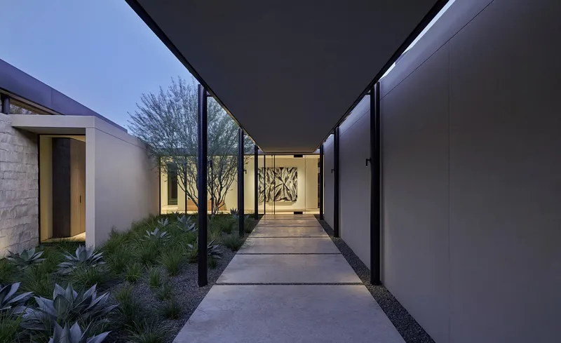 Covered outdoor entry passage with floating concrete walkway between planted courtyards at dusk
