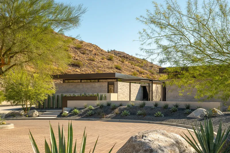Front facade with stone walls and flat roofs amid desert planting and rocky hillside