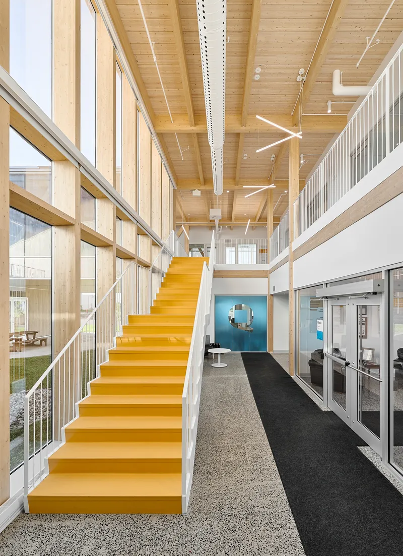 Double-height lobby with yellow steel stair and timber ceiling under linear light fixtures
