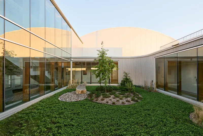 Planted courtyard with circular groundcover bed and trees surrounded by glass-walled corridors under an overcast sky