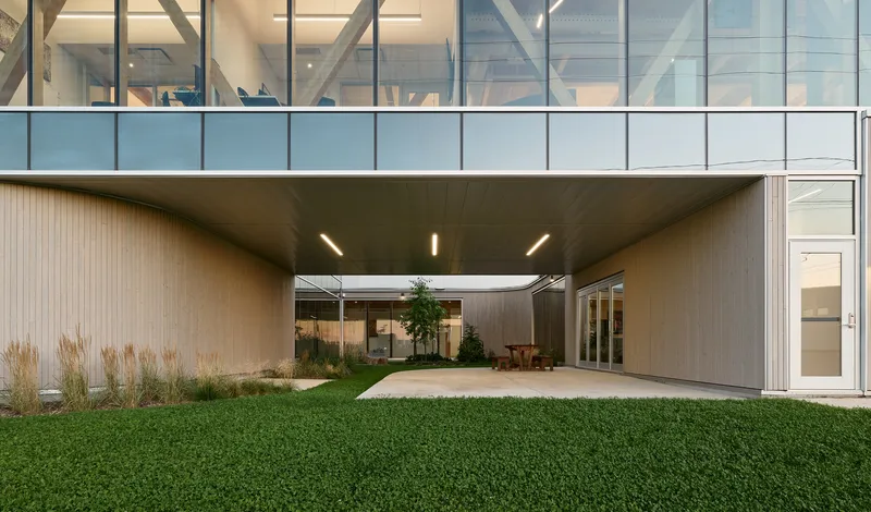 Covered passage with timber ceiling and linear lighting connecting glazed volumes across a lawn courtyard