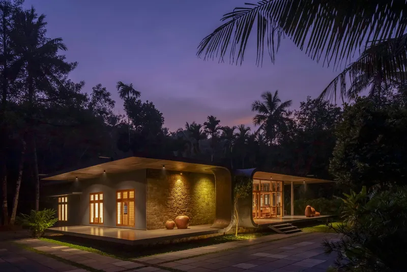 Curved roofline with illuminated stone wall and glazed entrance at dusk beneath palm trees