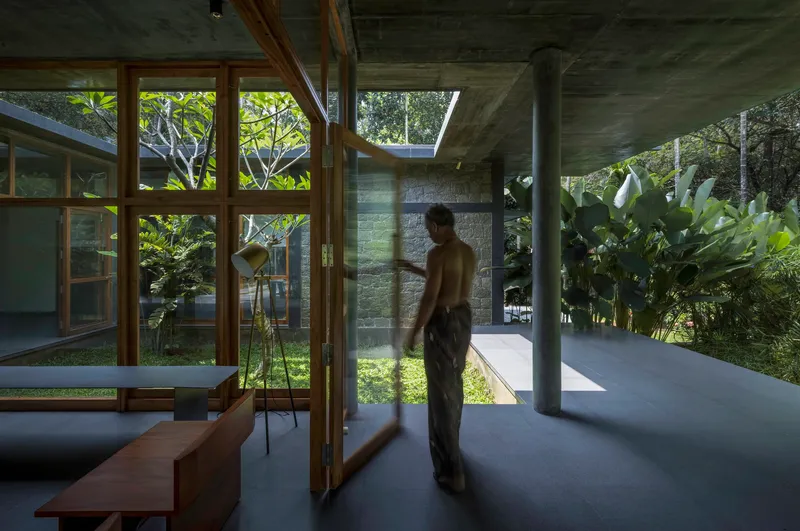 Open interior living space with board-formed concrete columns, timber glazing, and a figure passing through dappled sunlight