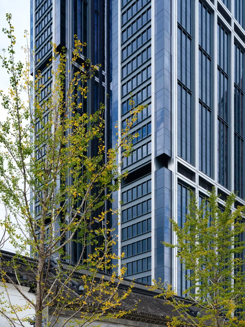 Glass tower facade with projecting bays behind trees with yellow autumn leaves and traditional tiled roof