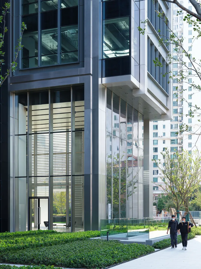 Ground-level lobby entrance with glass walls, horizontal louvers, and pedestrians walking on pathway through landscaped hedges
