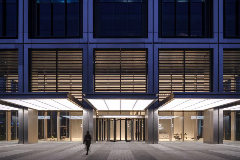 Tower entrance with illuminated canopy, revolving doors, and horizontal louvers at dusk with a pedestrian passing