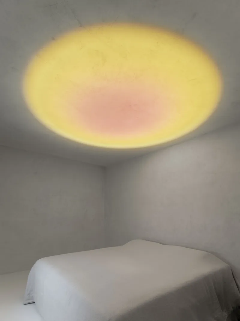 Corner of a white room with a circular skylight casting yellow and pink glow onto the ceiling
