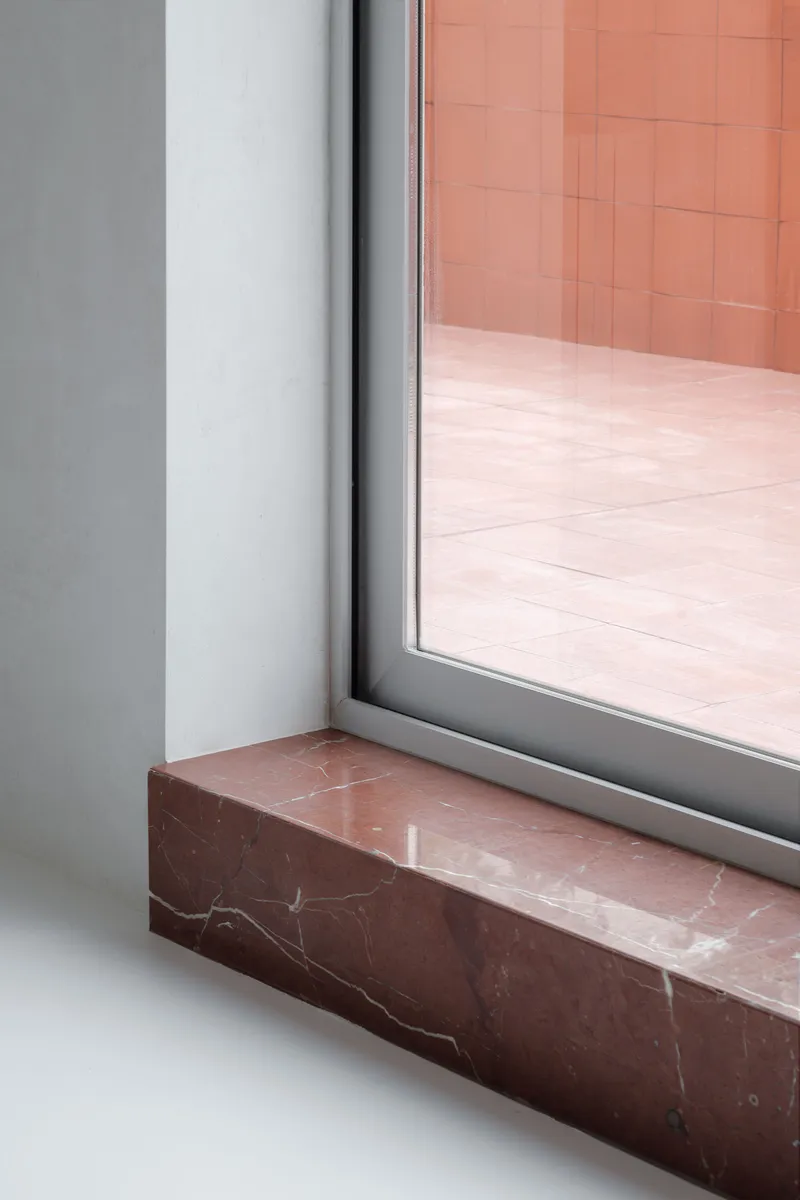 Detail of a window sill in red marble meeting white plaster walls and grey aluminium frame