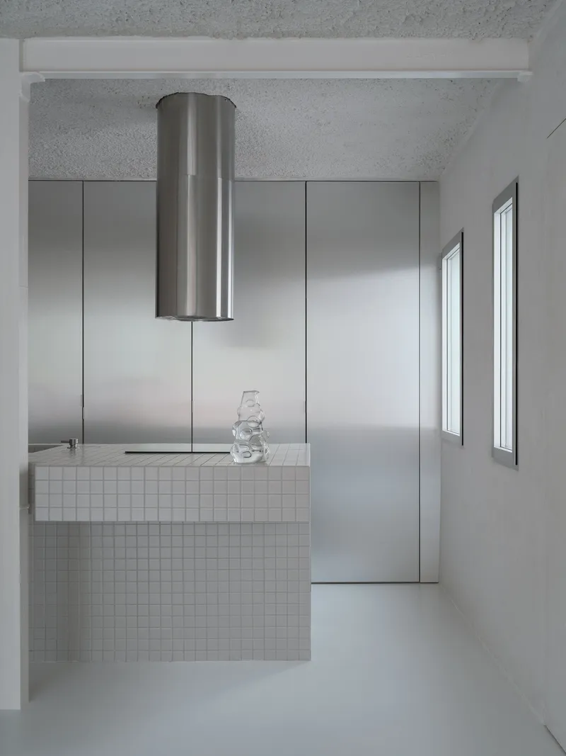 Kitchen island clad in white mosaic tile with cylindrical steel extractor above and glass vessel