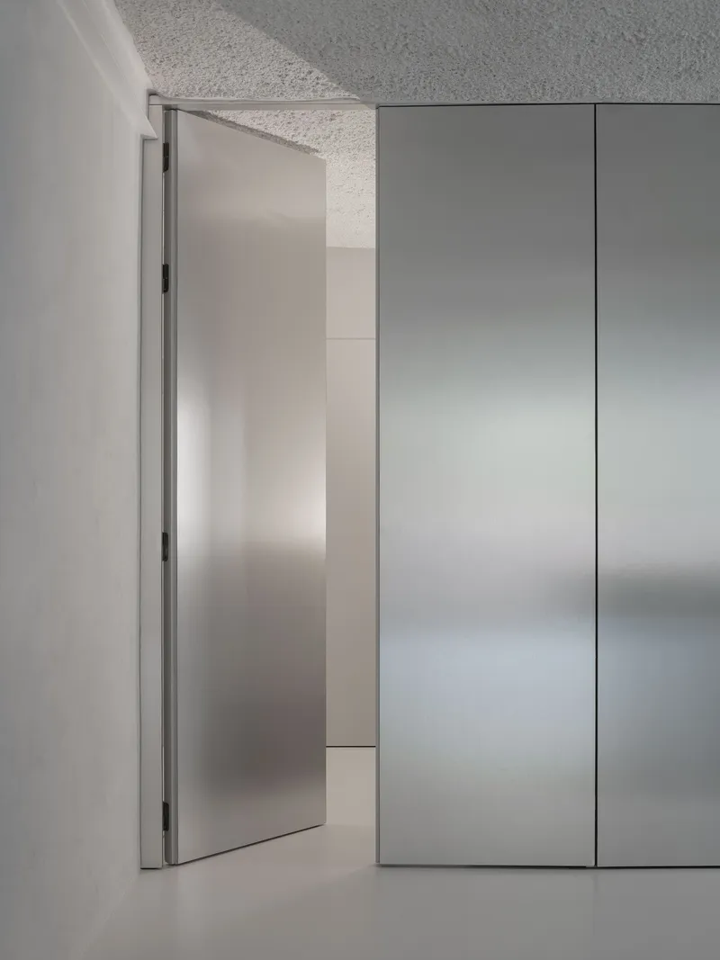 Brushed steel pivot door opening against floor-to-ceiling cabinet panels in soft grey light