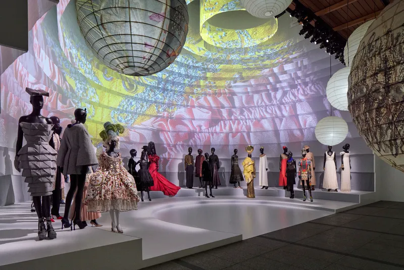 Gallery space with suspended lanterns and projected imagery across white stepped platforms holding fashion displays
