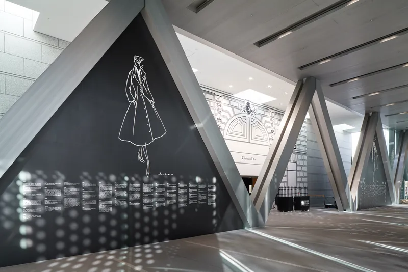 Exhibition corridor with diagonal concrete columns framing a black display wall and ornate facade beyond