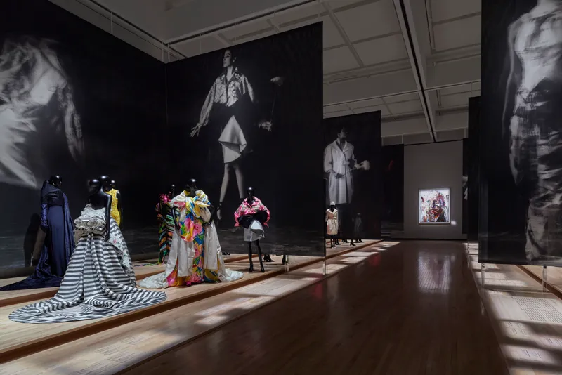 Exhibition room with dark walls showing projected images above mannequins and garments on wooden floor