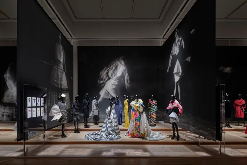 Gallery room with illuminated mannequins in colorful garments beneath large-scale black-and-white portrait projections