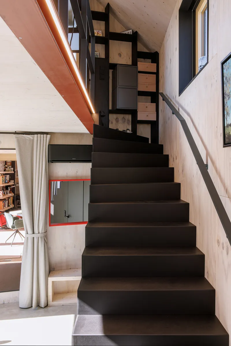 Black steel staircase with open risers ascending through a double-height space with light timber cladding