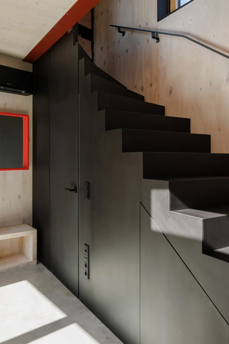 Black steel stair with integrated storage cabinets beneath treads against pale timber wall in afternoon light