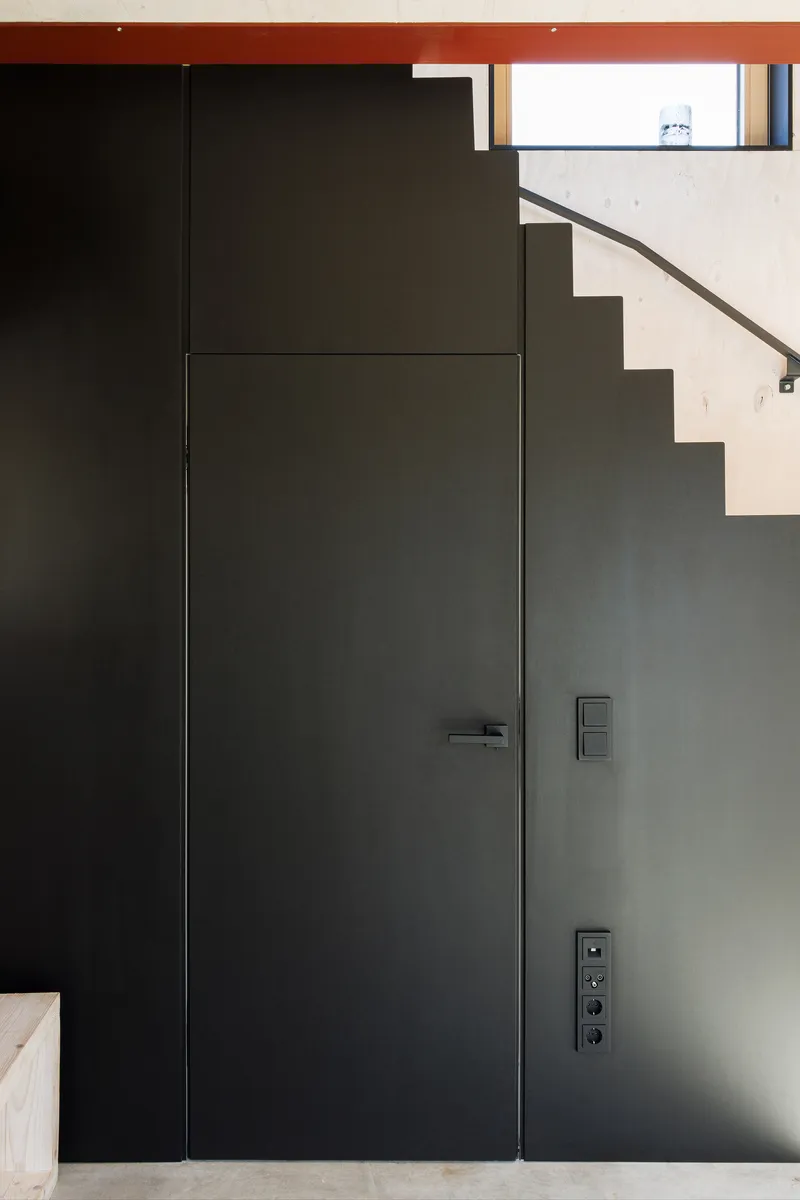Flush black metal door set into grey wall beneath visible stair treads and red ceiling beam