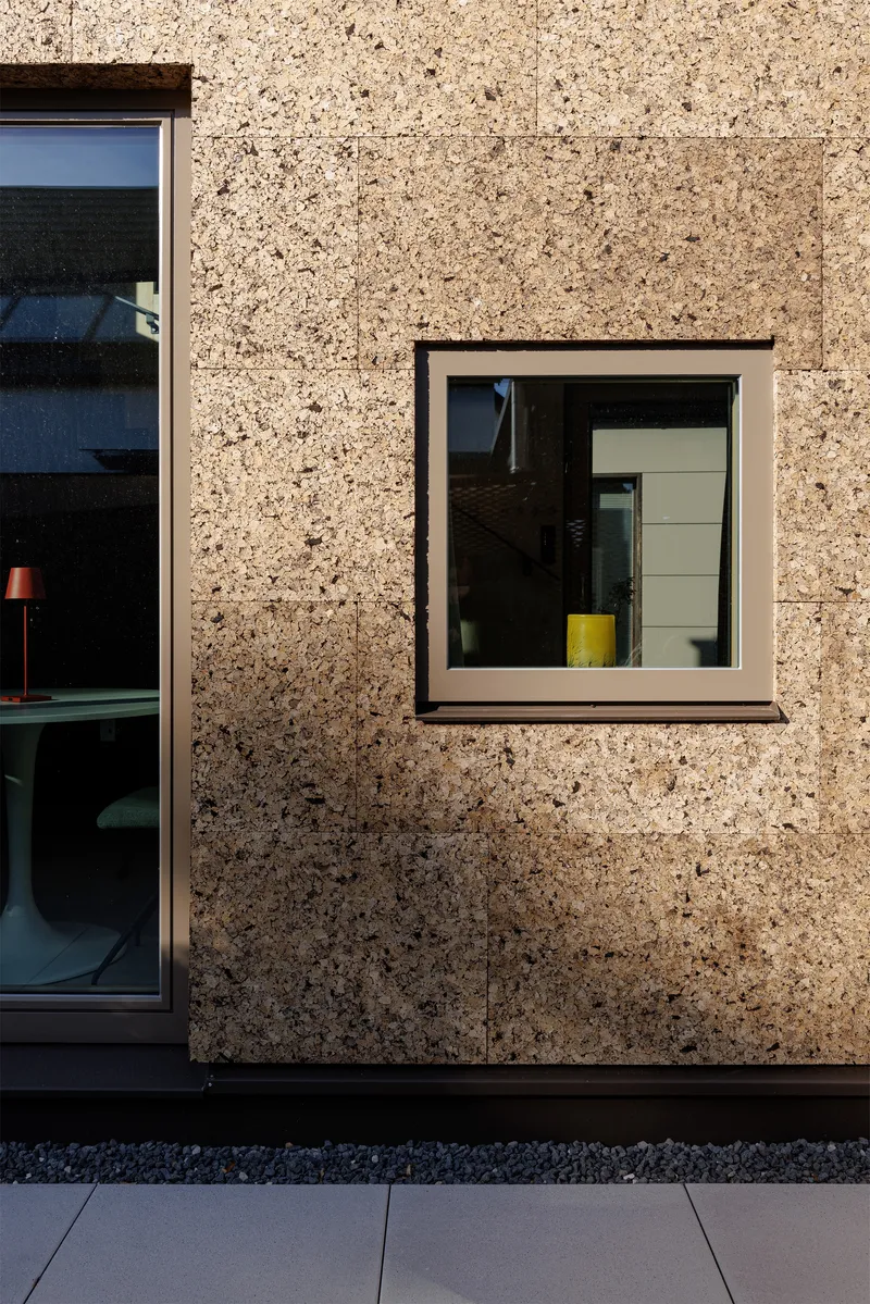 Cork aggregate facade with two aluminum-framed windows revealing interior spaces and a yellow stool