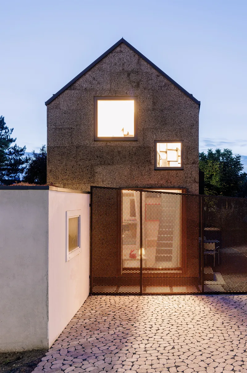 Gable-fronted cork facade with illuminated windows and perforated metal sliding doors at dusk