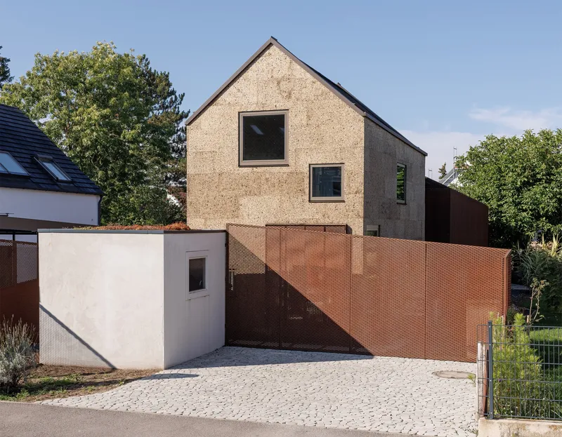 Street view of the cork-clad gable facade with perforated copper gate and white stucco entry volume