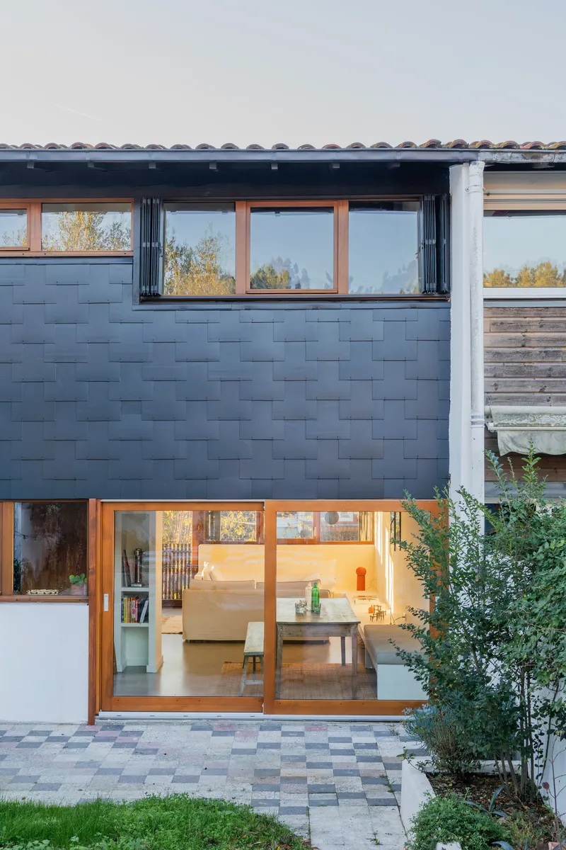 Rear elevation showing dark shingle wall with timber sliding doors opening to a paved courtyard at dusk