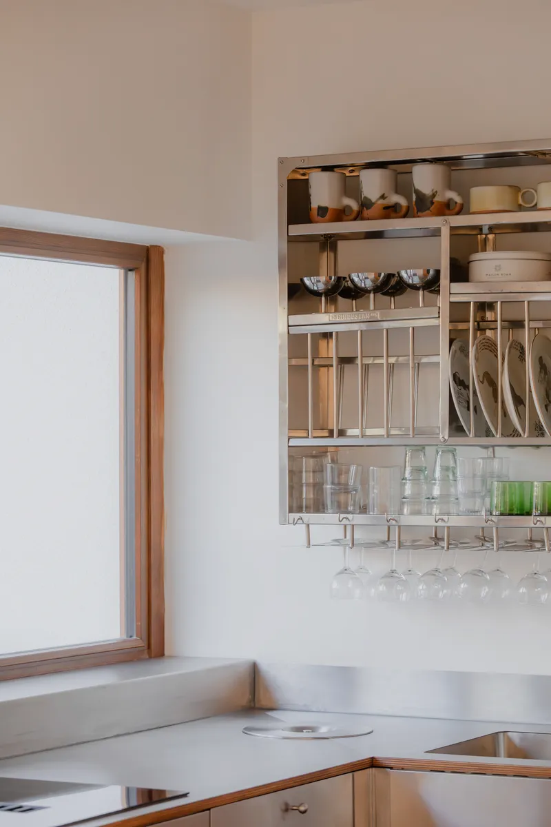 Kitchen corner with open steel shelving holding cookware, glassware and utensils beside timber-framed window