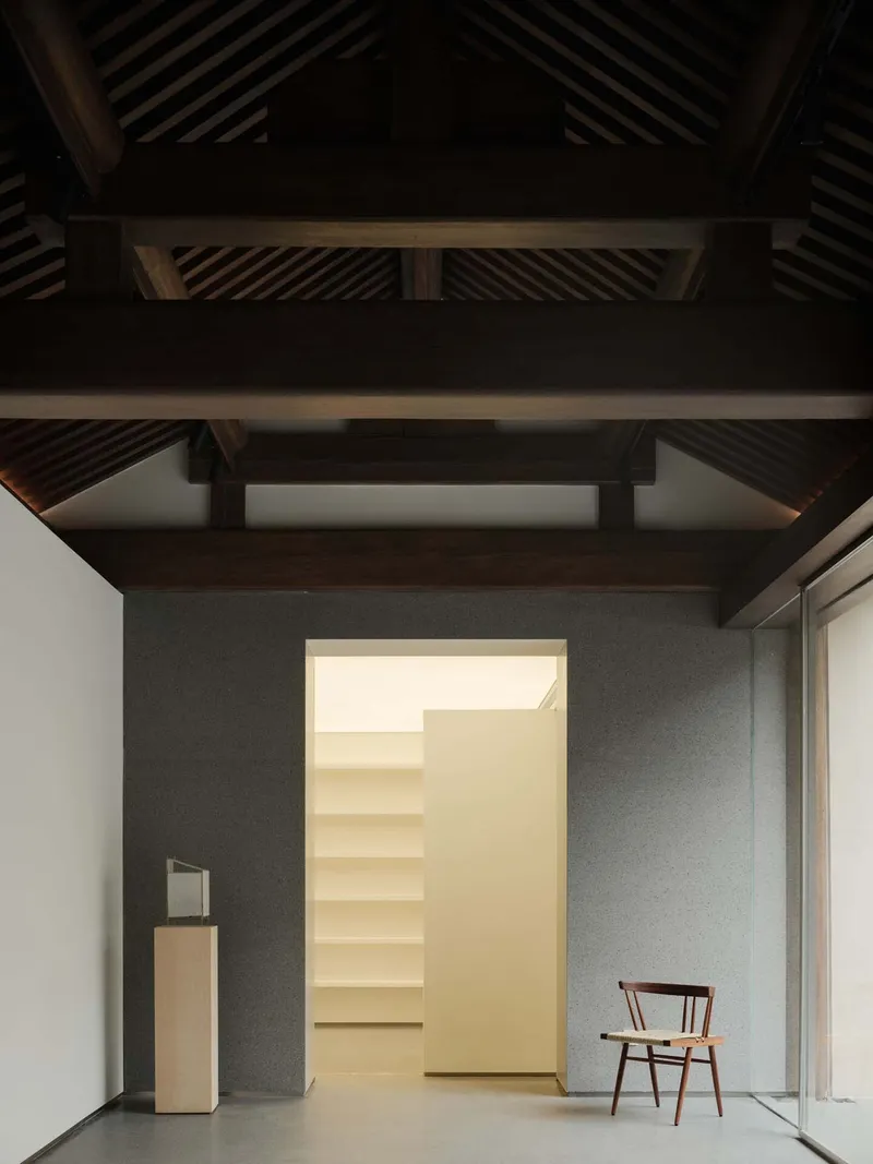 Exposed timber beam ceiling above grey walls framing a doorway into illuminated room with built-in shelving