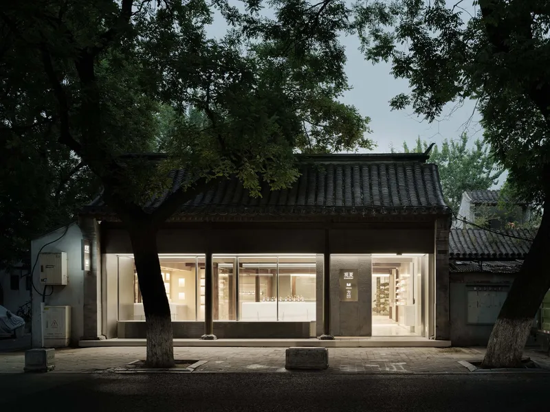 Night view of the illuminated glazed facade beneath traditional eaves with mature trees flanking the entrance