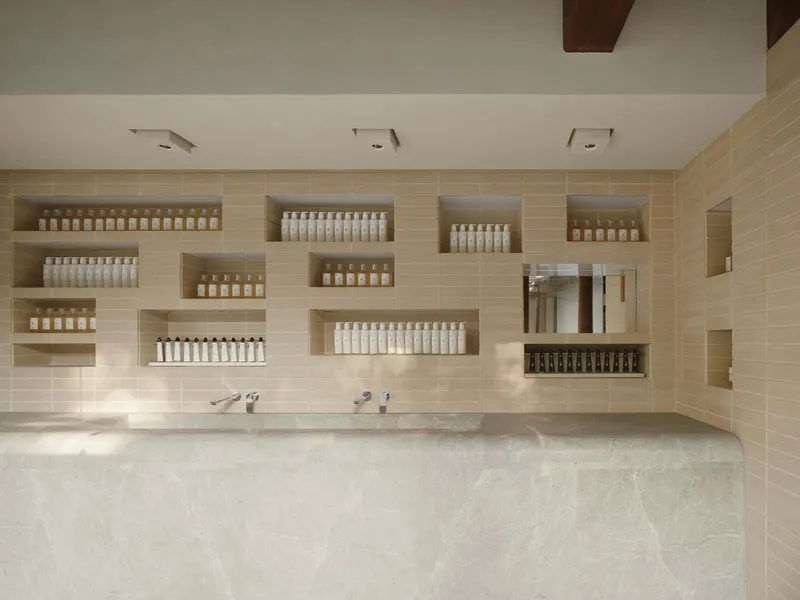 This architectural image showcases a modern bathroom interior with a minimalist design. The space features a beige tile wall with recessed shelves displaying va