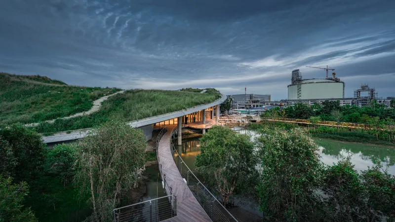 Elevated walkway bridging a planted wetland and pond at dusk with industrial tanks in the distance