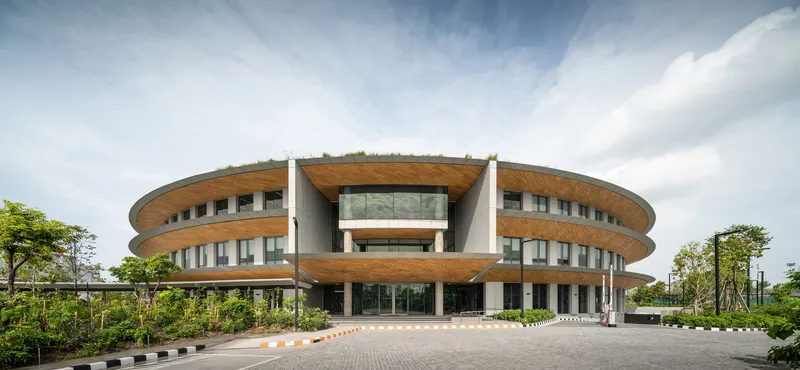 Front facade showing circular timber-clad levels with central glazed entrance canopy under cloudy skies