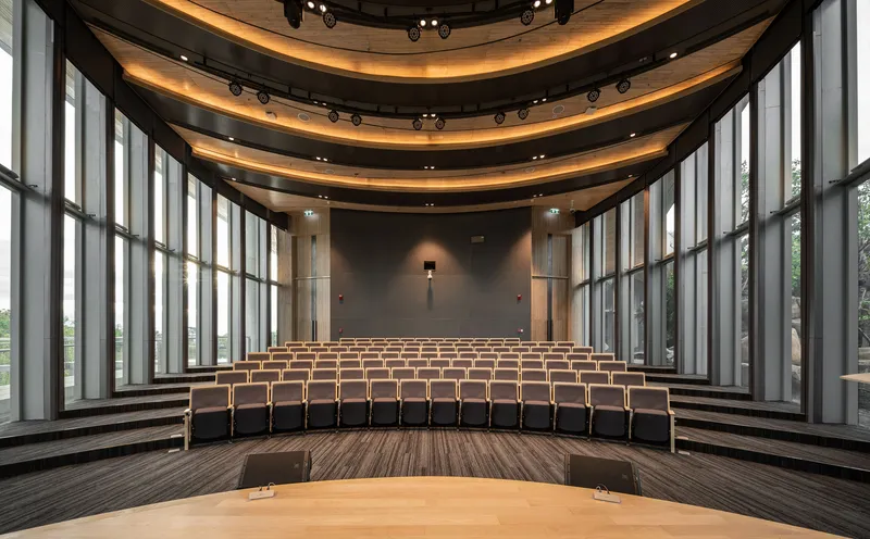 Auditorium with tiered wooden seating facing a stage framed by vertical glass walls