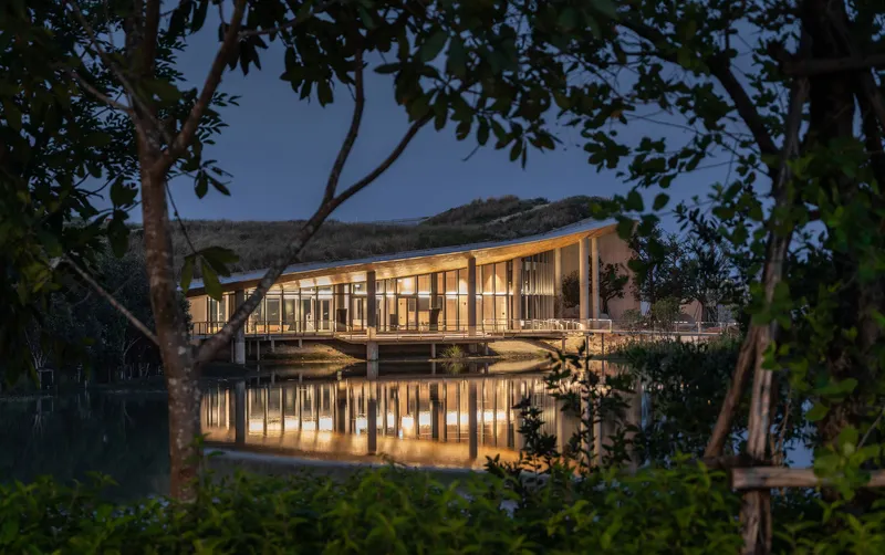 Glass pavilion on stilts above a lake reflecting interior lights at dusk