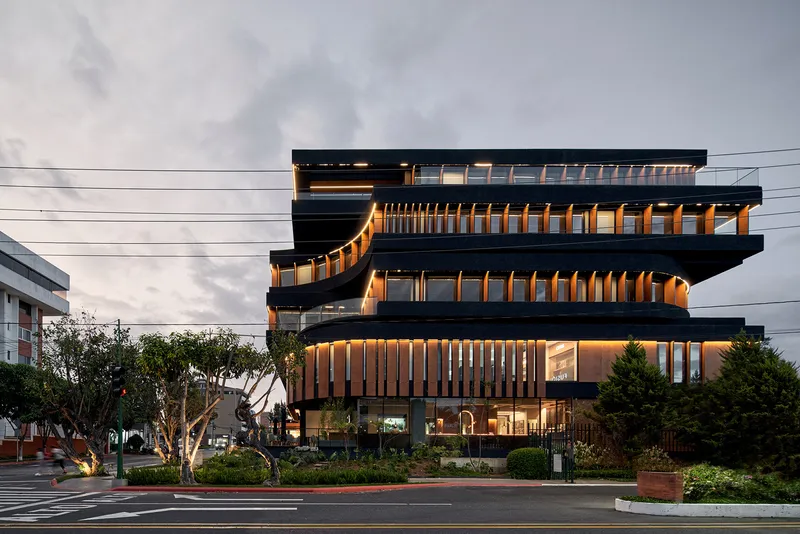 Layered facade with timber screening and dark concrete bands illuminated by warm interior light at twilight