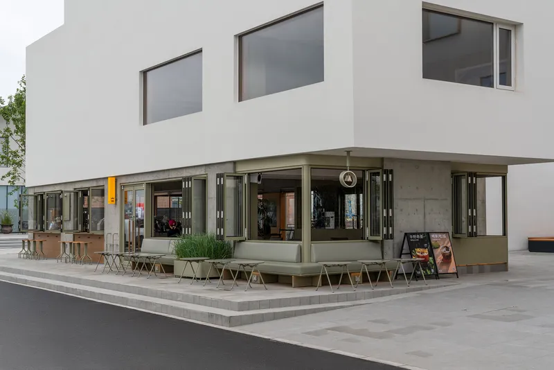 Corner detail of the folding glass storefront with olive-green frames and outdoor seating on the plaza