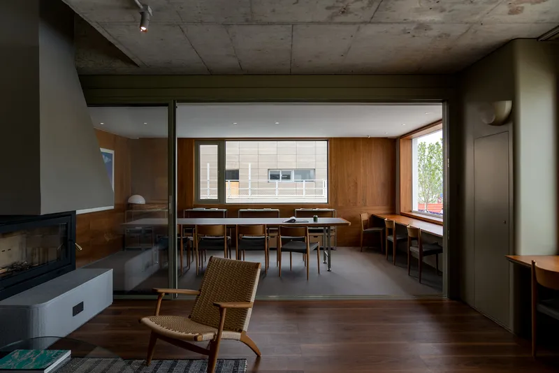 Interior view through wood-paneled spaces with exposed concrete ceiling and woven chair in foreground