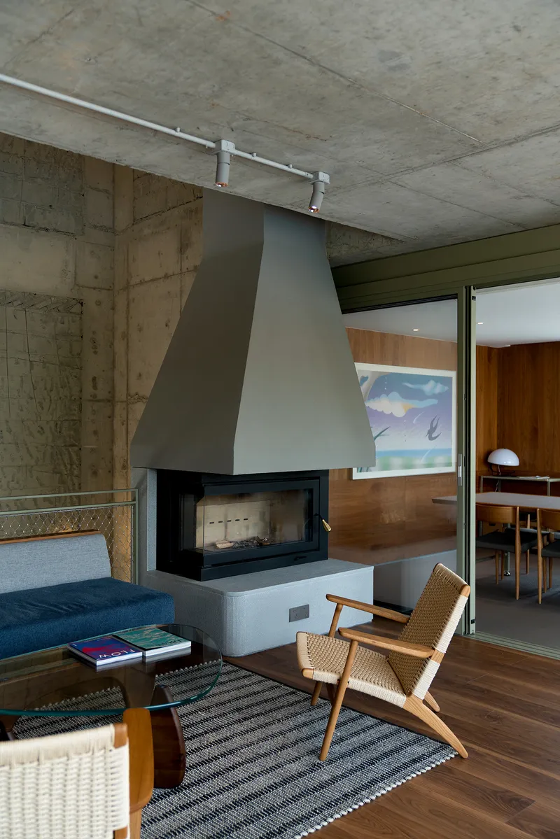 Living area with suspended metal fireplace hood, board-formed concrete walls, and woven lounge chair