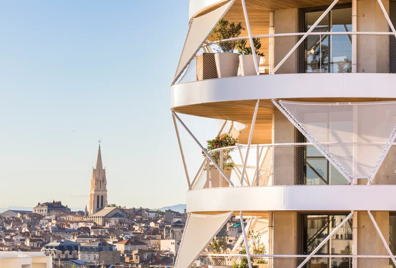 Stacked balconies with white diagonal structural bracing and planted terraces overlooking the city roofscape