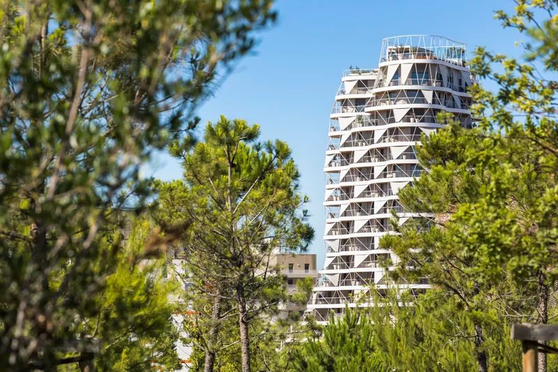 White residential tower with triangular balcony bracing rising above dense green treetops under clear sky