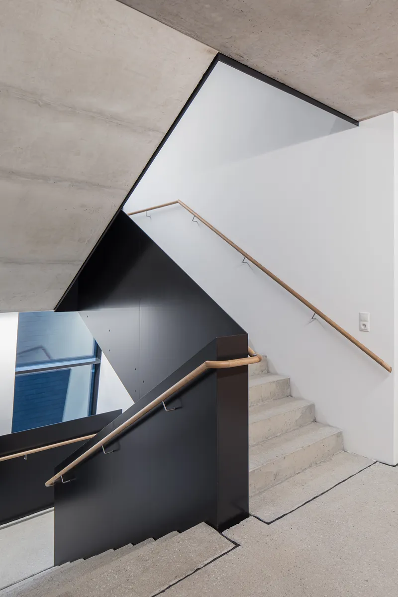Concrete stairwell with wood handrails and black steel balustrades under an angled skylight